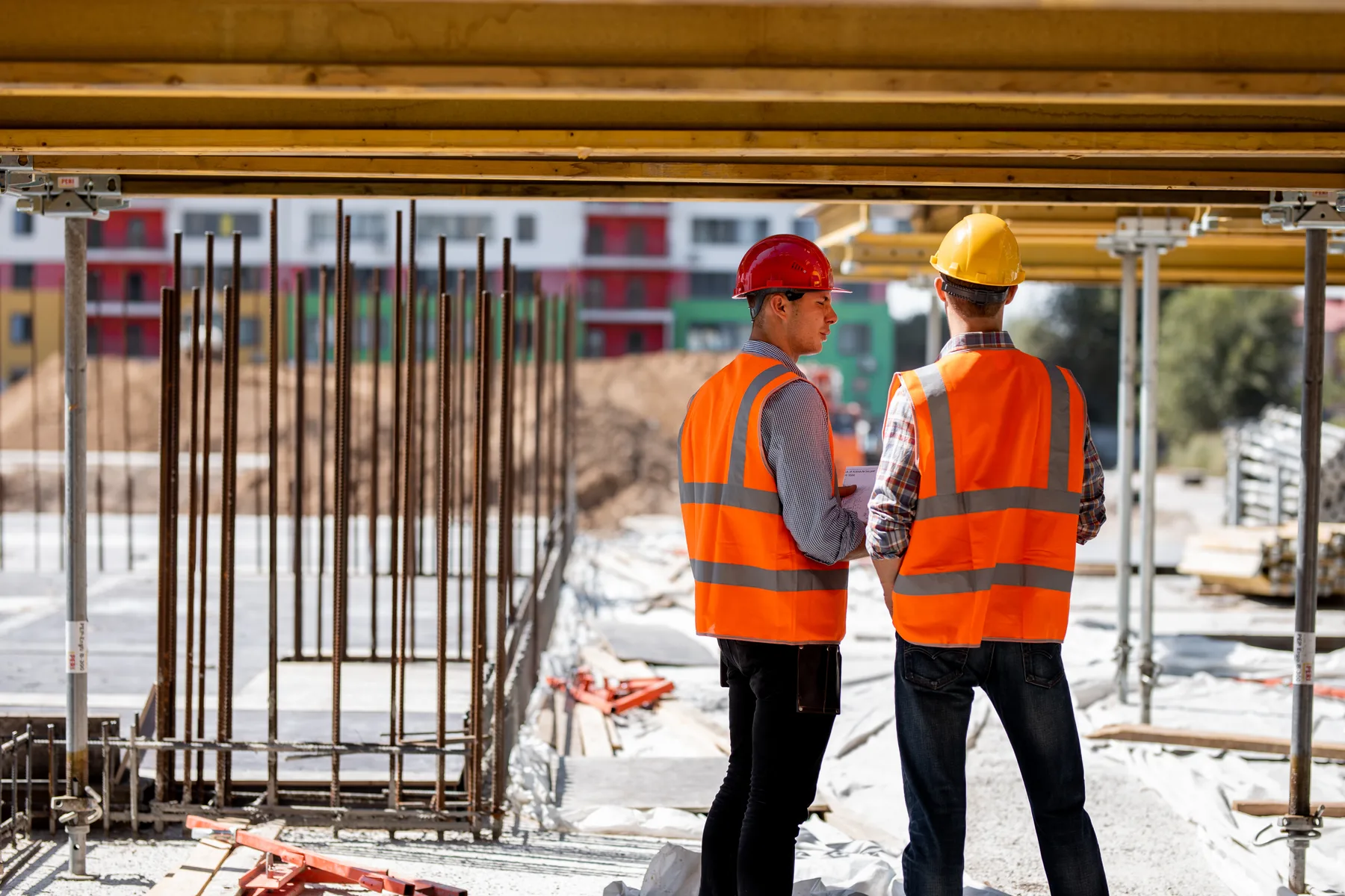 Two site leads in hard hats and orange vests talking under a formwork deck on a commercial jobsite