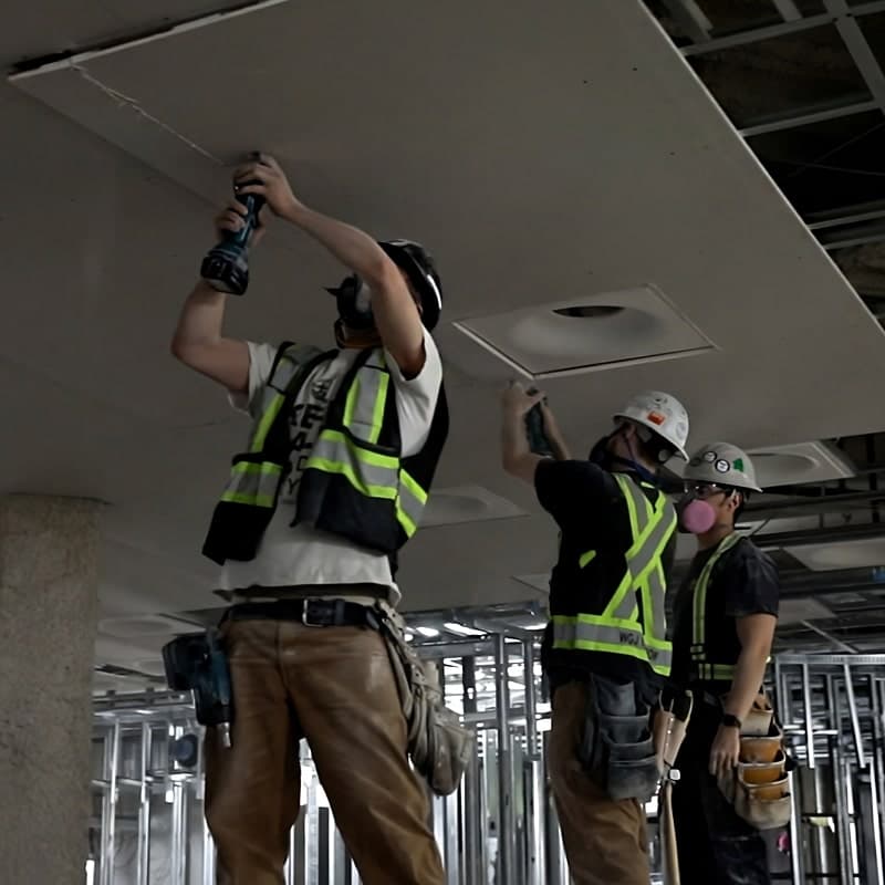 Three drywallers in hi-vis vests and hard hats finishing a ceiling, one driving screws overhead while the other two work the seams