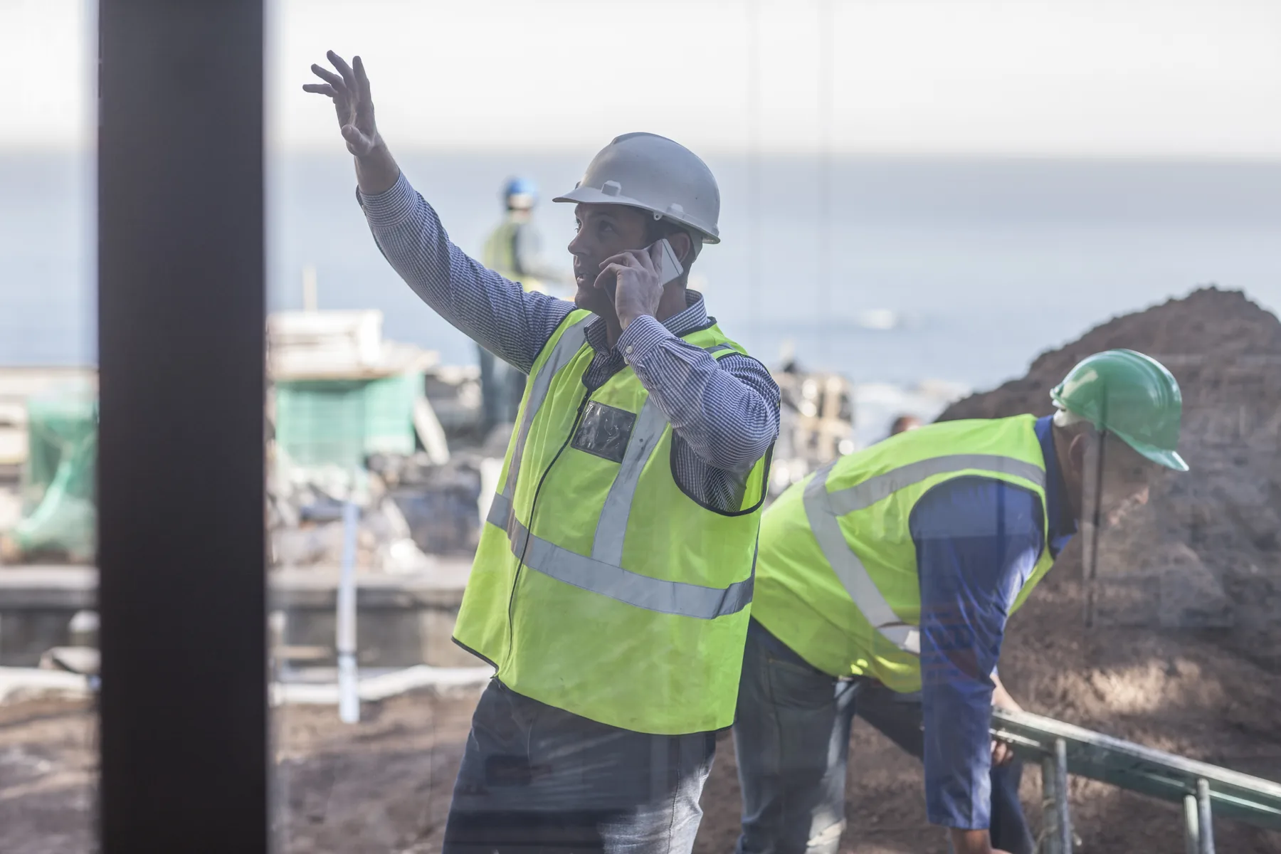 A site manager in a hard hat and hi-vis vest on a phone call, waving to direct work on a jobsite while a coworker checks a rail behind him