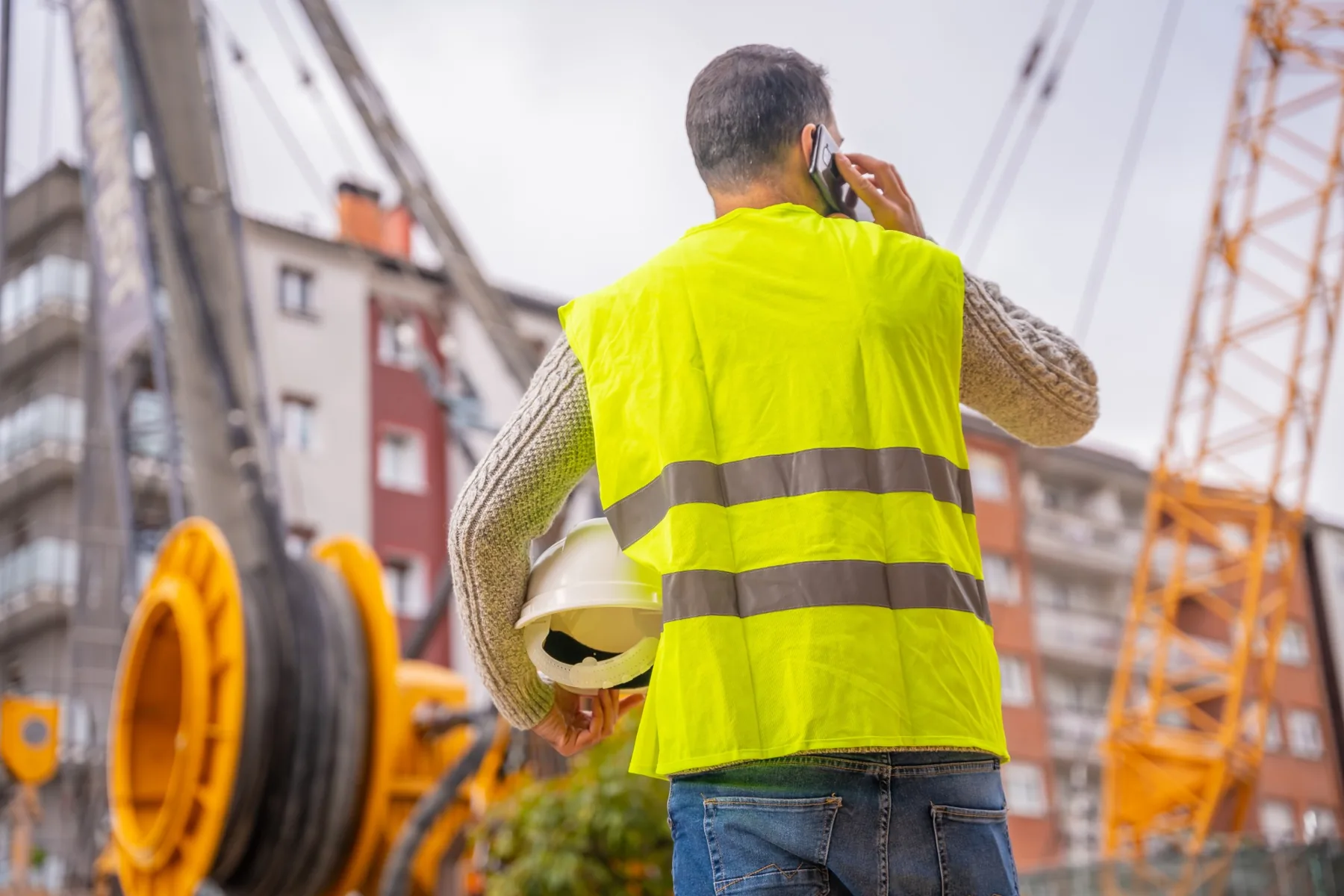 A site supervisor in a hi-vis vest, hard hat tucked under one arm, on a phone call on a jobsite with a crawler crane and cable reel in the background