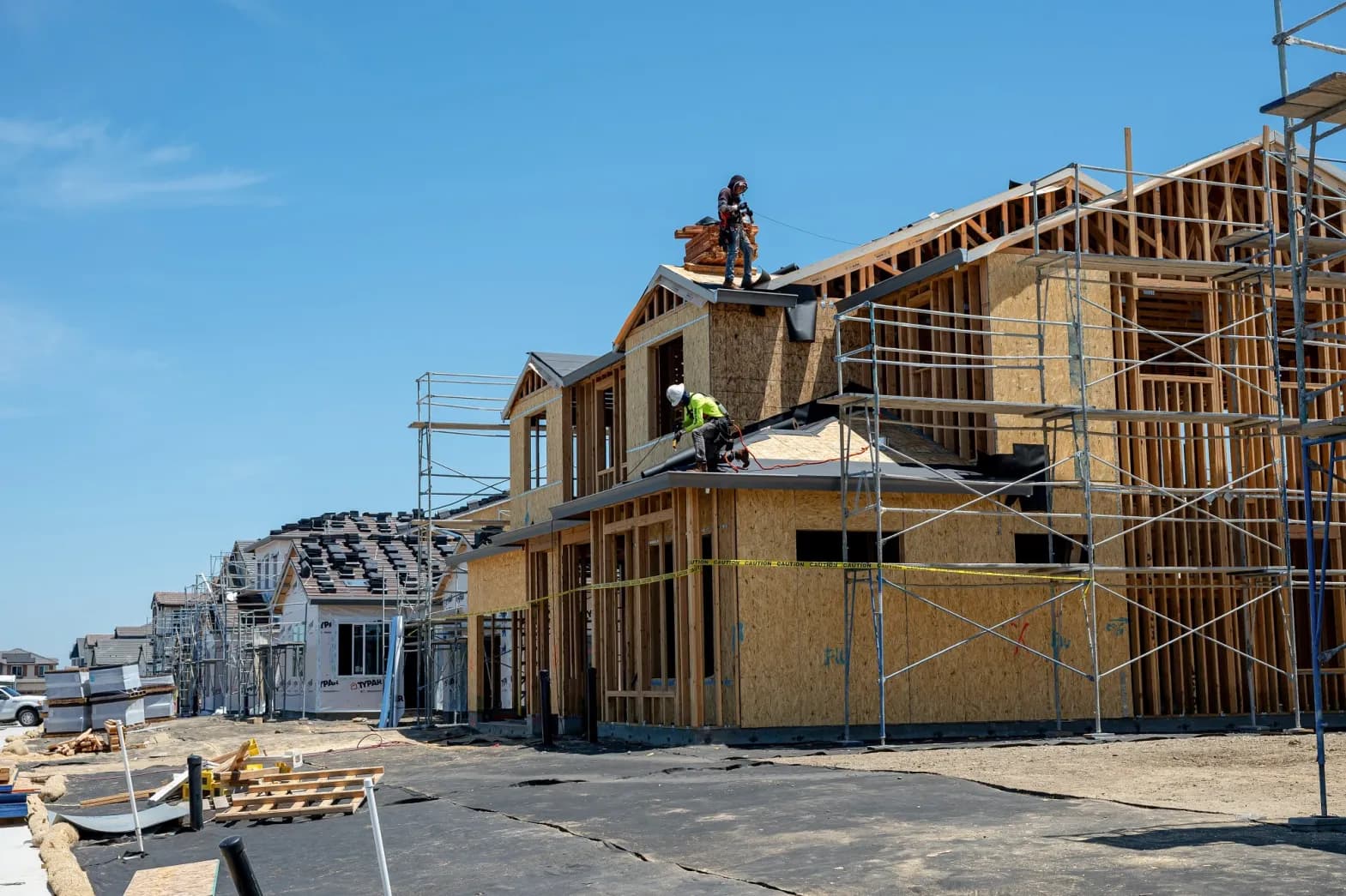 A row of houses under construction, framed and sheathed with scaffolding up, two workers on the roof of the nearest one