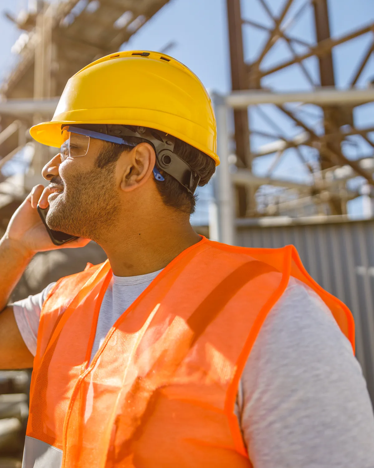 A construction engineer in a hard hat and hi-vis vest taking a phone call on a jobsite, steel structure behind him