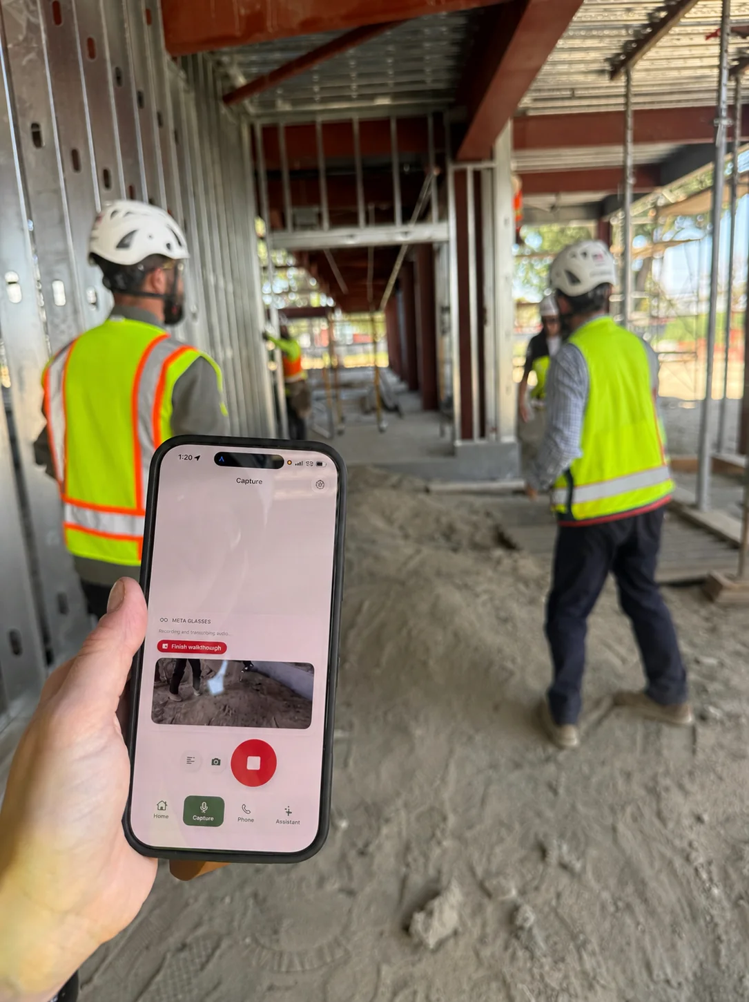 A superintendent holding a phone with Hardline recording a walkthrough, two workers in hi-vis vests moving through a steel-framed building behind them