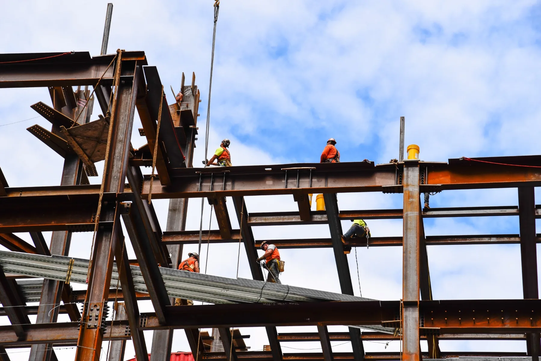 Ironworkers in hard hats and harnesses walking the steel beams of a building frame under construction, with metal decking bundles staged on the beams below