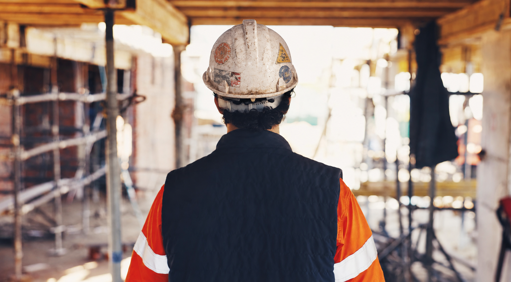 A construction superintendent in a scuffed hard hat and hi-vis jacket walking a site under formwork and scaffolding, the kind of site walk Hardline turns into daily logs, punch items, and tasks.