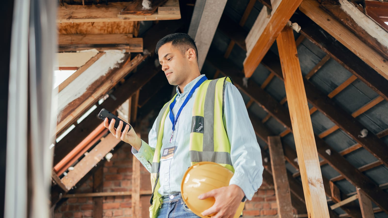 A construction engineer in a hi-vis vest checking his phone on a jobsite walk through a timber-framed roof, hard hat under his arm