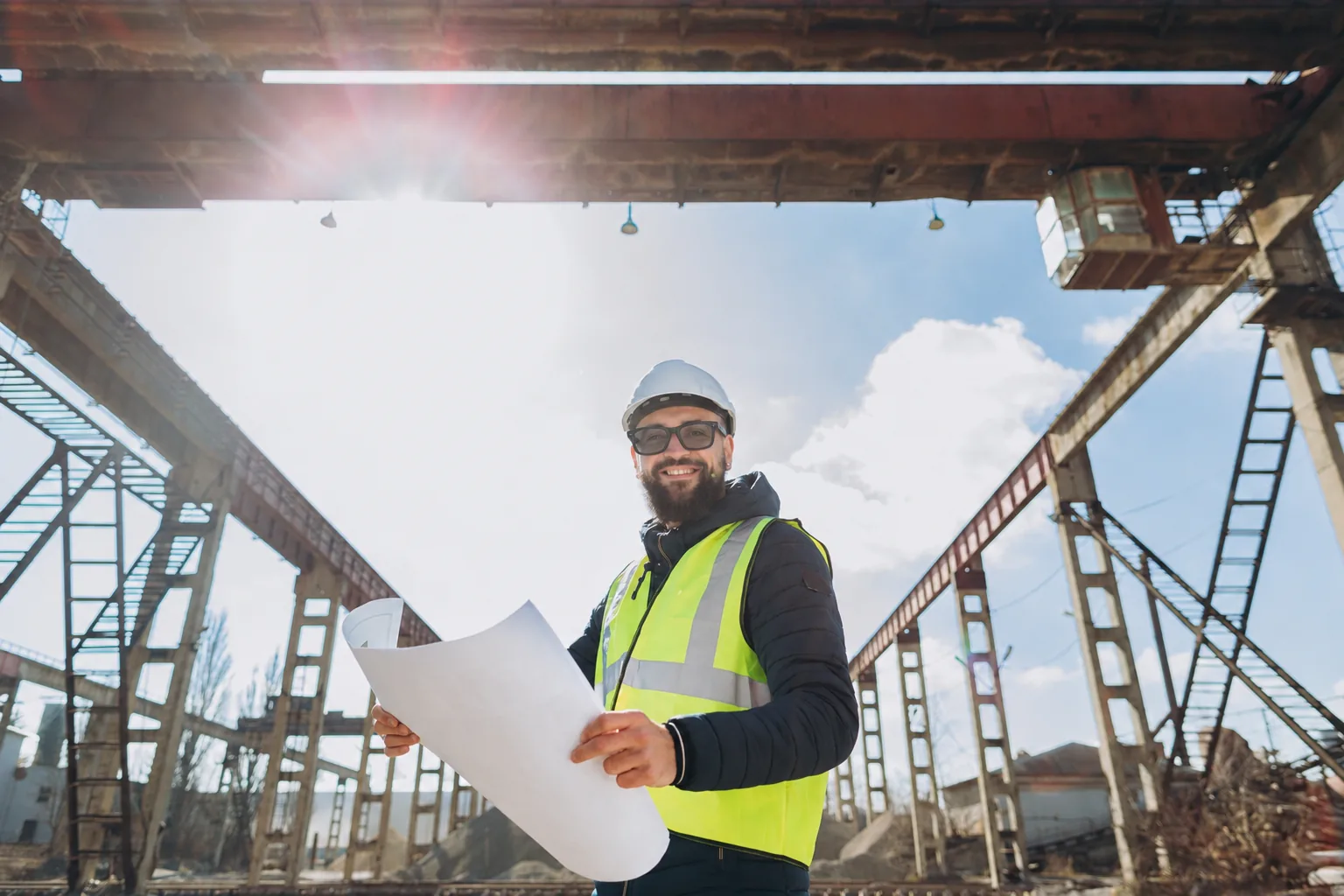A construction worker in a hard hat, safety glasses and hi-vis vest reviewing drawings on a steel-frame jobsite, where voice AI and smart glasses have to fit the work.