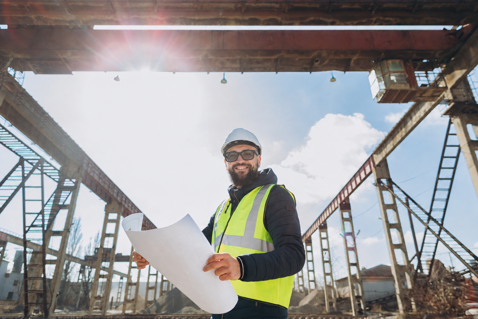 Construction worker in a hard hat, safety glasses, and hi-vis vest reviewing drawings on a steel-frame commercial job site, the environment where smart glasses have to prove themselves.